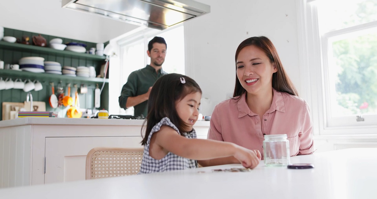 A young girl and her mother play with pretend money at the kitchen table, while her father watches in the background