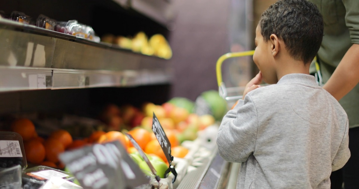 A child at the grocery store observes fresh produce while shopping with a guardian, learning about real-world budgeting